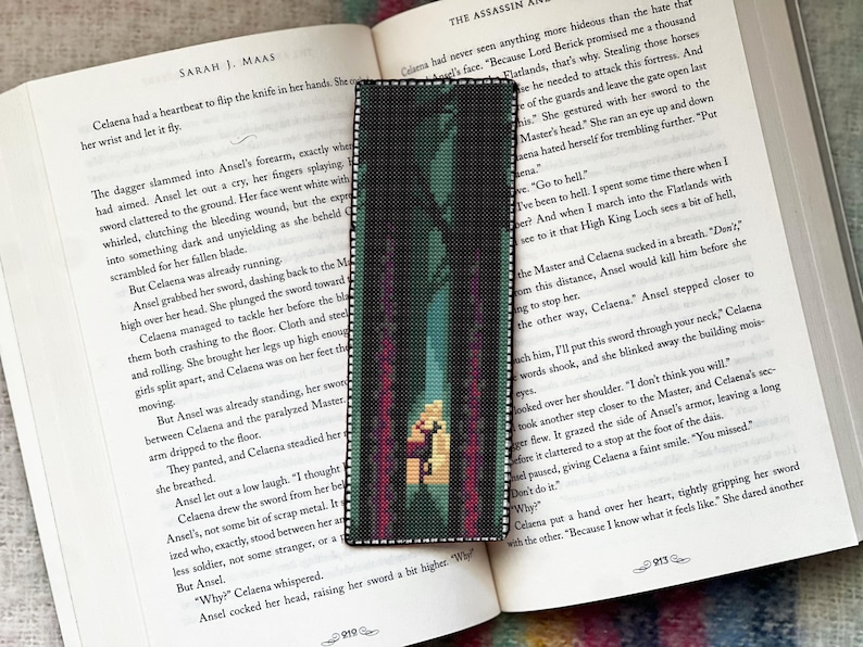 May include: A cross-stitch bookmark depicting a forest scene with a central figure. The bookmark is rectangular and rests on an open book with text by "Sarah J. Maas". The book is open to page 919.