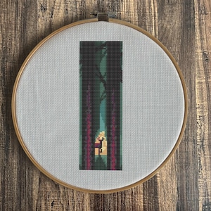 May include: A cross-stitch embroidery in a wooden hoop depicts a pixelated forest scene. The design features tall, dark trees framing a path leading to a light source. The color palette includes greens, purples, and a warm yellow glow.