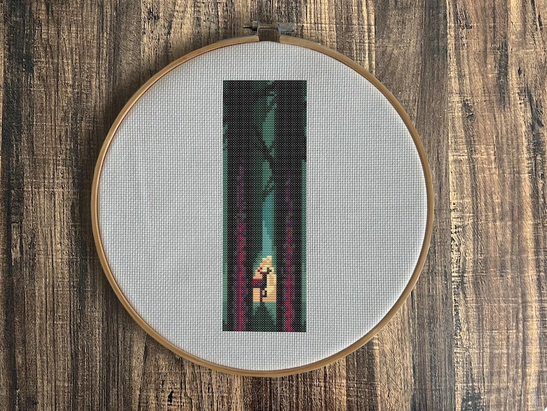 May include: A cross-stitch embroidery in a wooden hoop shows a pixelated forest scene. The design features tall, dark trees framing a path leading to a light source. The colour palette includes greens, purples, and a warm yellow glow.