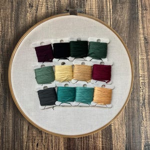 May include: An embroidery hoop with white fabric and a grid pattern. Twelve spools of colourful embroidery floss are arranged in rows. Colours include dark green, teal, yellow, and burgundy. The hoop is on a wooden surface.