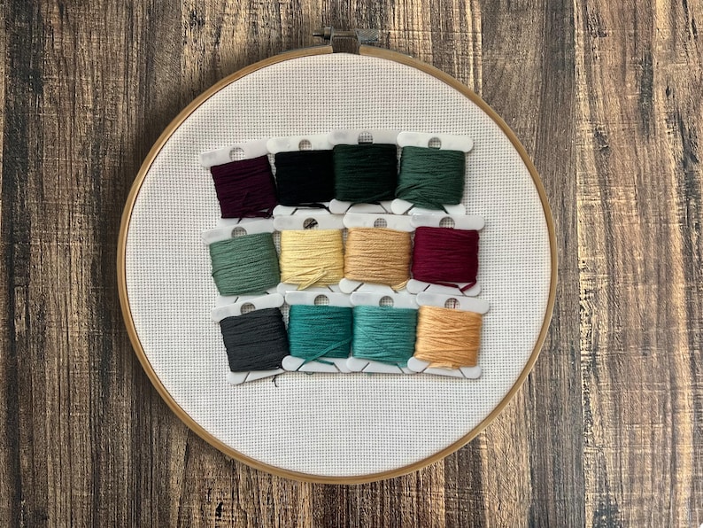 May include: An embroidery hoop with white fabric and a grid pattern. Twelve spools of colourful embroidery floss are arranged in rows. Colours include dark green, teal, yellow, and burgundy. The hoop is on a wooden surface.