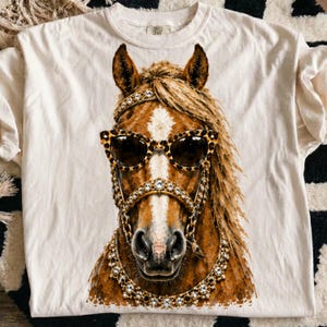 May include: A cream-colored t-shirt featuring a graphic print of a brown horse wearing leopard-print sunglasses and a gold-toned bridle adorned with clear crystals. The horse has a prominent white blaze on its face and a textured mane.