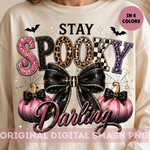 Könnte beinhalten: Ein cremefarbenes Sweatshirt mit dem Schriftzug STAY SPOOKY in verschiedenen Mustern wie rosa Glitzer, Schwarz, Leopardenmuster und Karos. Eine große schwarze Schleife befindet sich in der Mitte über zwei rosa Kürbissen und dem Wort Darling.