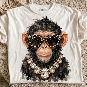 Puede incluir: Una camiseta color hueso con una ilustración gráfica de un chimpancé que lleva gafas de sol con estampado de leopardo y un gran collar de cristal transparente con múltiples facetas. El mono tiene pelaje oscuro y rasgos detallados.