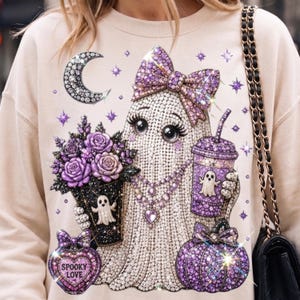May include: A beige sweatshirt featuring a graphic of a ghost adorned with a purple bow, holding a bouquet of purple roses and a purple drink cup. The design includes a crescent moon, a heart-shaped perfume bottle labeled Spooky Love, and a pumpkin.