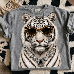 Könnte beinhalten: Ein graues T-Shirt mit Rundhalsausschnitt und einem großen Grafikdruck eines weißen Tigers, der eine Sonnenbrille mit Leopardenmuster und eine silberfarbene Halskette mit einem großen, rechteckigen schwarzen Edelsteinanhänger trägt.