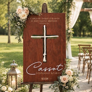 May include: A wooden wedding sign on an easel, featuring a cross made of rope. The sign has the text "A CORD OF THREE STRANDS IS NOT EASILY BROKEN" and the names "Cassot, Stephanie & Grant" with the date 5/2/26. Floral arrangements adorn the sign.