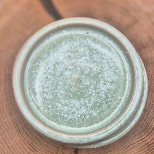 May include: Three ceramic plates stacked together, featuring a light green glaze with a speckled effect. The plates have a circular form with a raised rim and a brown edge. They are placed on a wooden surface.