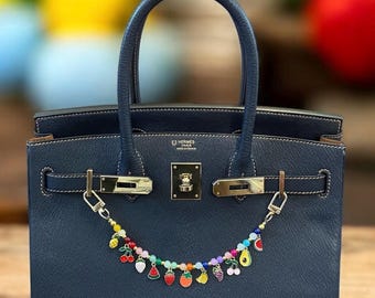 Könnte beinhalten: Eine dunkelblaue Ledertasche mit goldenen Beschlägen und einem Charm-Armband mit bunten Frucht-Anhängern. Das Charm-Armband ist am Schloss der Handtasche befestigt.