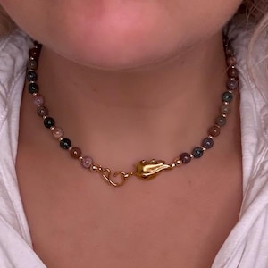 Op de afbeelding: Een ketting met een reeks veelkleurige, ronde edelstenen in tinten bruin, groen en roze. Het heeft een goudkleurige sluiting en een decoratieve goudkleurige hanger. De ketting is ontworpen om om de nek te worden gedragen.