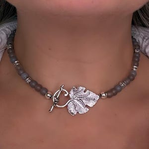May include: A gray beaded necklace with a silver leaf pendant and toggle clasp. The necklace features small, round gray beads and decorative silver accents. The leaf pendant is detailed with intricate veining.