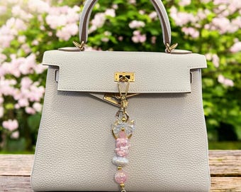 Puede incluir: Un bolso de mano de cuero gris claro con asa superior y herrajes dorados. Un charm decorativo con cuentas rosas y blancas y un colgante de corazón cuelga del cierre. El fondo presenta flores rosas borrosas.