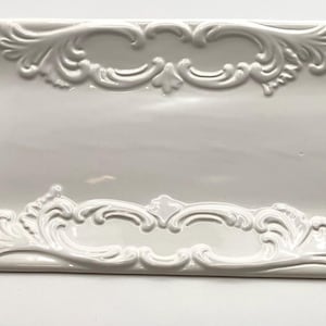 May include: A rectangular white ceramic serving tray featuring a raised, ornate scrollwork border along all four edges. The center of the tray is smooth and flat, while the edges are decorated with intricate, symmetrical relief patterns.