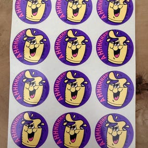 May include: A sheet of twelve circular stickers, each featuring a cartoon character resembling a yellow cup with a purple liquid splashing out. The character has a wide, happy expression and the text AHHHHHH! is printed in pink along the left side.