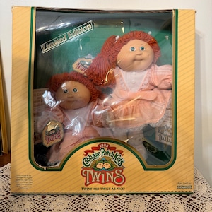 May include: A 1985 Cabbage Patch Kids Twins limited edition doll set in original packaging. Two dolls with red yarn hair and green eyes wear matching light pink dresses with white lace trim. The box features the text Cabbage Patch Kids TWINS.