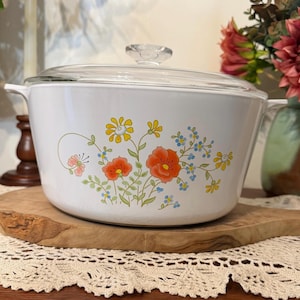 Peut inclure: Une cocotte blanche avec un couvercle en verre transparent, ornée d'un motif floral composé de fleurs orange, jaunes et bleues avec des tiges et des feuilles vertes sur le devant. La marmite possède deux poignées latérales et un bouton en verre.
