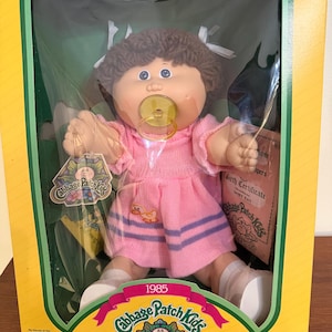 Puede incluir: Muñeca Cabbage Patch Kids vintage en su embalaje original. La muñeca tiene cabello castaño, ojos azules y un chupete. Viste un vestido rosa con un aplique de pato y rayas moradas. La caja es amarilla y verde, con el logo de Cabbage Patch Kids y el texto "1985".