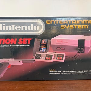 May include: A Nintendo Entertainment System Action Set box. The box is black with the Nintendo logo and the words "ACTION SET" in red. Includes a console, two controllers, a Zapper light gun, and a double game pak with Duck Hunt and Super Mario Bros.