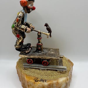 May include: A detailed figurine of a clown operating a small train car, with a bird perched on a lever. The clown is dressed in a colorful outfit with a red nose and orange hair. The base is a stone with gold bead accents.