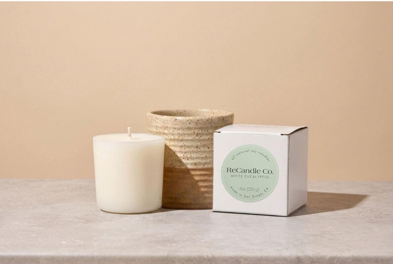 A white eucalyptus scented soy wax candle refill placed next to it’s box and a handmade, ceramic refillable candle jar