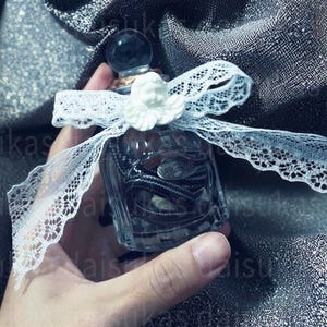 Puede incluir: Un frasco de perfume de vidrio transparente con una serpiente negra y blanca en su interior, coronado con un orbe negro y un querubín blanco. La botella está atada con una cinta de encaje blanco.