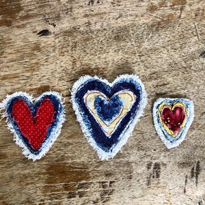 May include: Three heart-shaped fabric patches with frayed white edges. The left patch is red with white dots, the center patch features layered blue denim and yellow fabric, and the right patch displays a small red heart with yellow and blue borders.