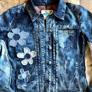 May include: A blue acid-wash denim jacket featuring a pointed collar, a button-front closure, and a single chest pocket. The front is decorated with three fabric flower appliqués in varying shades of blue, and the sleeves have light-colored panels.