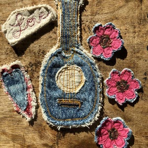May include: A collection of denim fabric patches featuring a guitar shape with yellow stitching, three pink flower-shaped patches with dark centers, a heart-shaped denim patch with red accents, and a white fabric patch with the word Love stitched in red.