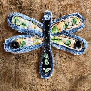 May include: A dragonfly-shaped fabric applique featuring frayed blue denim edges. The wings are crafted from a yellow and white floral patterned fabric with green accents. The central body is black with white dots and green stitched details.