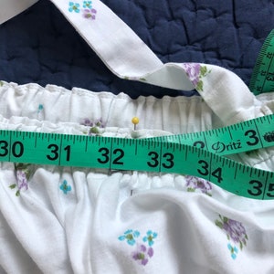 Pu&ograve; includere: Tessuto bianco con un motivo floreale di fiori blu e viola. Un metro a nastro verde mostra una misura da 76 a 89 cm. Uno spillo giallo &egrave; inserito nel tessuto. Il tessuto ha una vita elastica arricciata e spalline.