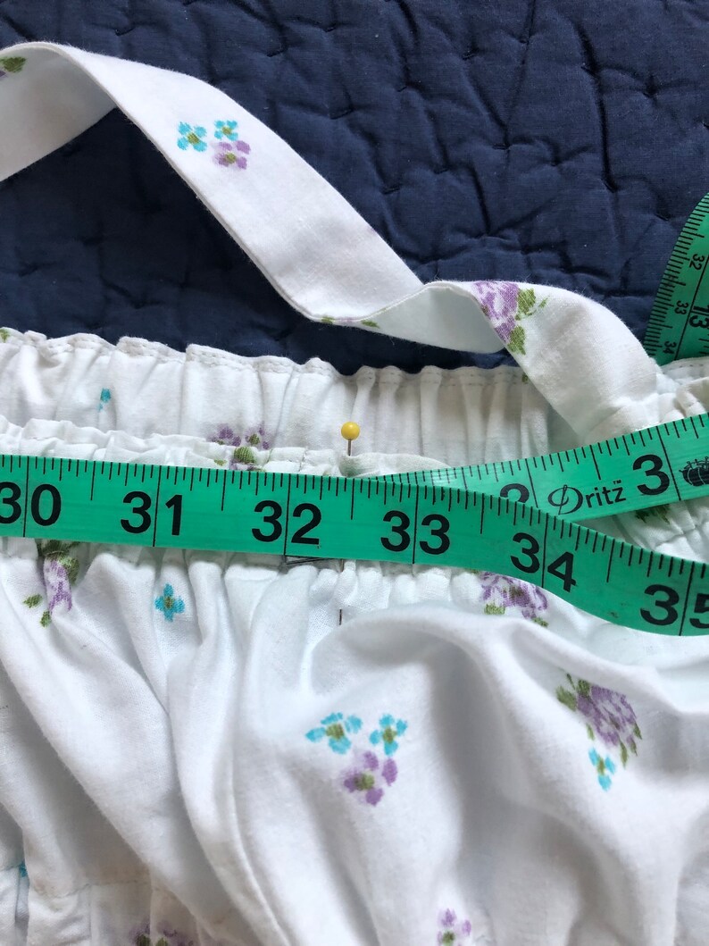 Pu&ograve; includere: Tessuto bianco con un motivo floreale di fiori blu e viola. Un metro a nastro verde mostra una misura da 76 a 89 cm. Uno spillo giallo &egrave; inserito nel tessuto. Il tessuto ha una vita elastica arricciata e spalline.