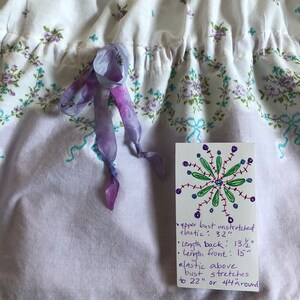 Pu&ograve; includere: Abito in tessuto bianco con motivo floreale viola e turchese. L'abito ha uno scollo a balze e un fiocco di nastro viola. L'etichetta recita: busto superiore non allungato: 81 cm, lunghezza schiena: 34 cm, lunghezza davanti: 38 cm.