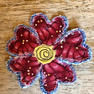 May include: A fabric flower applique featuring five heart-shaped petals in a deep red fabric with small yellow dots. The edges of the petals are frayed and blue, and the center is a circular yellow patch with a black stitched spiral design.