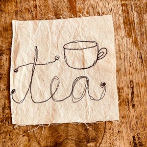 May include: A square piece of off-white fabric featuring black thread embroidery. The design consists of a simple line-drawn teacup positioned above the word tea, which is written in a continuous, cursive-style script with small circular knots.