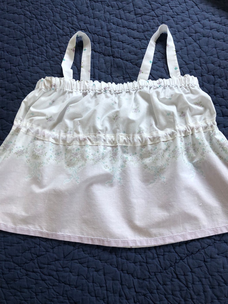 Pu&ograve; includere: Top a camisole bianco con spalline sottili e design a balze. Il top presenta un motivo floreale nei toni del viola e del verde. Le spalline sono decorate con lo stesso motivo floreale.