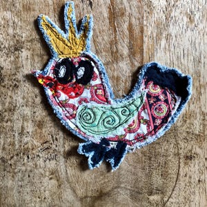 May include: A fabric applique shaped like a bird wearing a yellow crown. The bird features a white base with red and pink paisley patterns, a light green wing with dark green swirl embroidery, black eyes, and dark blue frayed fabric edges.