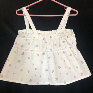 Pu&ograve; includere: Top a camisole bianco con un motivo floreale di piccoli fiori viola e verdi. Il top ha uno scollo arricciato e spalline, appeso a un gancio rosa. Il tessuto &egrave; leggero e sembra essere di cotone.