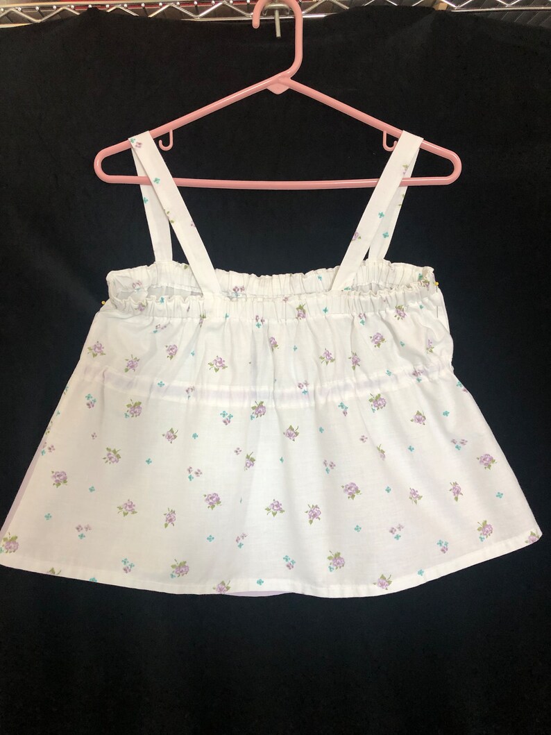 Pu&ograve; includere: Top a camisole bianco con un motivo floreale di piccoli fiori viola e verdi. Il top ha uno scollo arricciato e spalline, appeso a un gancio rosa. Il tessuto &egrave; leggero e sembra essere di cotone.