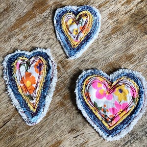 May include: Three heart-shaped fabric patches featuring layered denim and floral-patterned cotton fabric. Each patch has frayed white edges, yellow fabric borders, and visible red stitching securing the layers together in a decorative design.