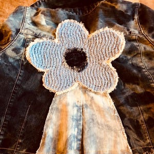 May include: A dark blue denim jacket featuring a large, light blue, textured fabric flower appliqué on the back. The flower has a dark center and frayed edges. Below the flower, a triangular, light-wash denim panel is stitched onto the jacket.