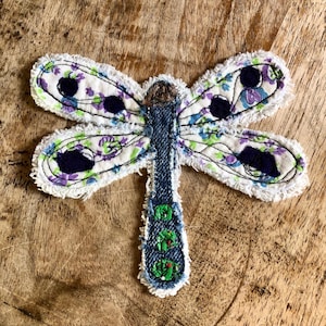 May include: A dragonfly-shaped fabric applique featuring a denim body with green embroidered circles and four wings made of white fabric with a purple and green floral pattern, dark blue circular accents, and frayed white edges.