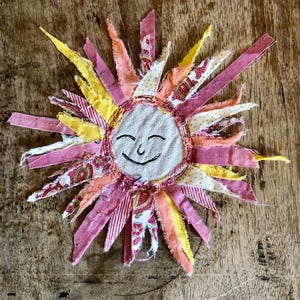 May include: A fabric sun ornament featuring a central circular face with embroidered eyes and a smile. The sun rays are made of various strips of fabric in shades of pink, yellow, orange, and white with floral, paisley, and polka dot patterns.