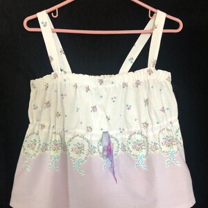 Pu&ograve; includere: Top camisole bianco con motivo floreale e a nastro. La parte superiore ha una sezione inferiore color lilla e sottili spalline. La parte superiore &egrave; appesa a una gruccia rosa su sfondo nero.