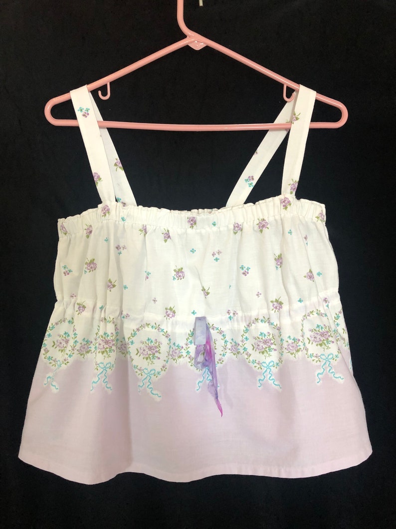 Pu&ograve; includere: Top camisole bianco con motivo floreale e a nastro. La parte superiore ha una sezione inferiore color lilla e sottili spalline. La parte superiore &egrave; appesa a una gruccia rosa su sfondo nero.