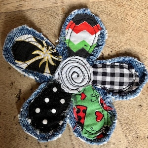 May include: A fabric flower applique featuring five petals with frayed denim edges. Each petal displays a different pattern: black with yellow starbursts, red-green-white chevron, black-white gingham, black with white polka dots, and green with ladybugs.