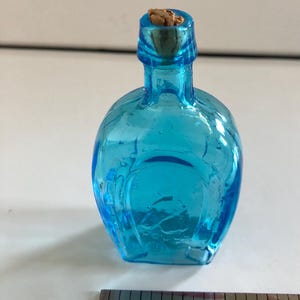 May include: A small, antique, turquoise-colored glass bottle with a cork stopper. The bottle has a rounded body and a narrow neck. The bottle is approximately 3 inches tall.