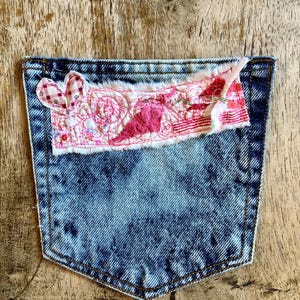 May include: A blue acid-wash denim pocket featuring a rectangular fabric patch stitched to the top. The patch is decorated with pink and red embroidery, a small heart-shaped fabric appliqué, and frayed edges with visible loose threads.