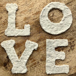 May include: Four individual fabric letters spelling the word LOVE, crafted from an off-white, quilted textile material. Each letter features a textured, diamond-stitched pattern and frayed, raw edges, presenting a distinct, tactile appearance.