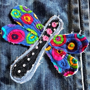 May include: A fabric dragonfly applique featuring four wings with a blue background and multicolored circular patterns in pink, green, and orange. The central body is black with white polka dots and pink accents, all stitched onto blue denim fabric.
