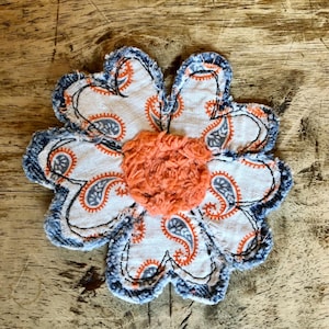 May include: A fabric flower applique featuring six petals made of white material with a grey and orange paisley pattern. The edges of the petals are finished with frayed blue denim fabric. A textured, fuzzy orange circle forms the center.